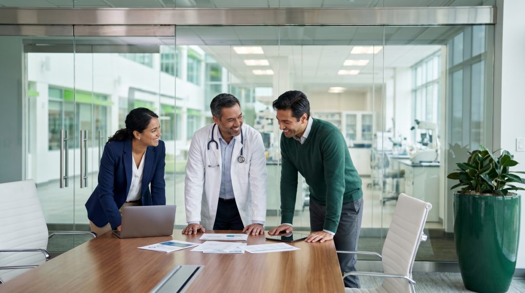 Een cross-functional pod-team in de farmaceutische industrie, bestaande uit een zakelijke professional, een arts in een witte jas en een productspecialist, overlegt glimlachend rond een vergadertafel met documenten en een laptop in een modern, glazen kantoor.