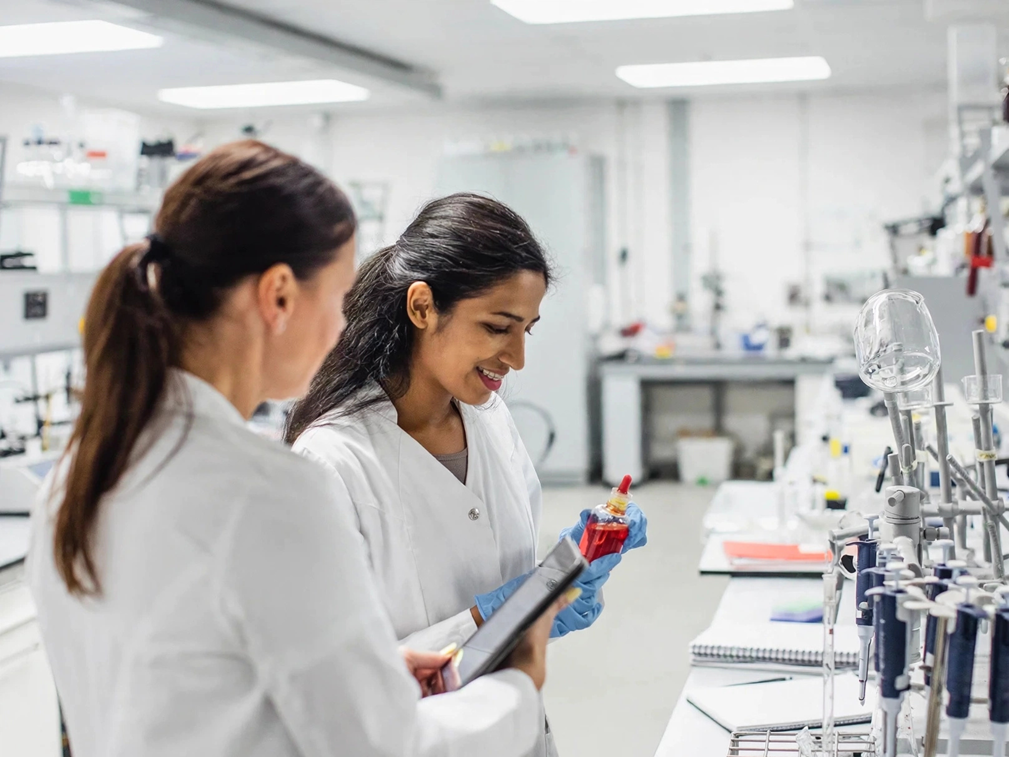 Twee vrouwen in witte jassen in een laboratorium, waarvan de rechter handschoenen draagt en een flesje met rode vloeistof en een tablet vasthoudt