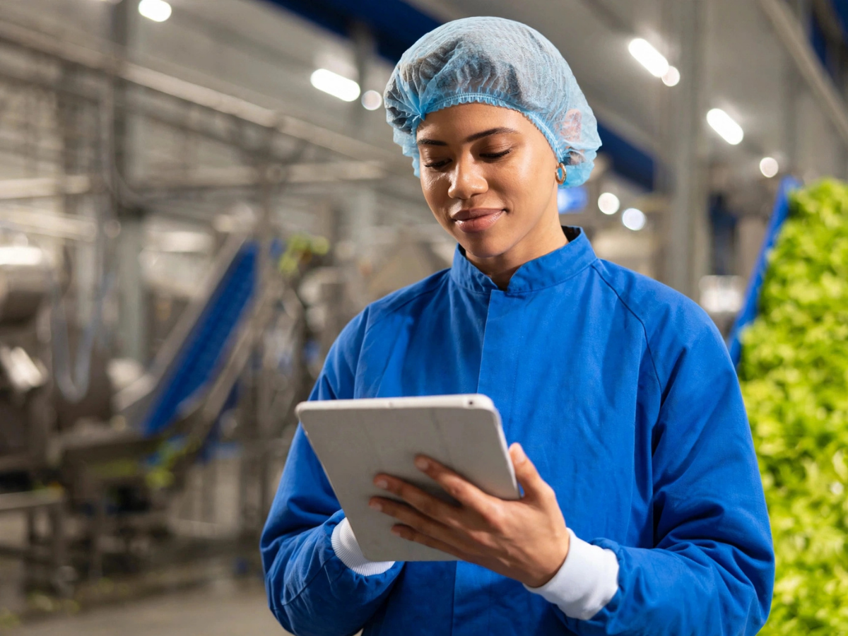 Kwaliteitscontroleur in blauwe hygiënekleding gebruikt een tablet in een food & beverage productiefabriek.